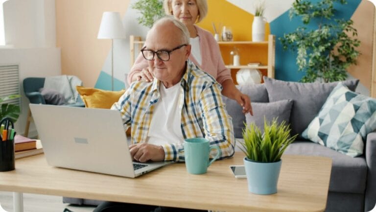 a senior man using laptop while his wife touching his shoulder looking at screen of the laptop