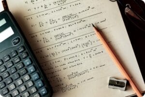 Close up photograph of an open ruled notebook filled with complex handwritten calculations, alongside a calculator, a wooden pencil and a metal sharpener