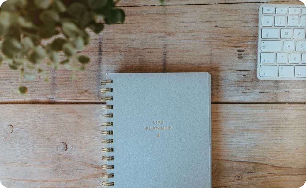 Top-down view of a light gray spiral-bound "Life Planner" notebook resting on a rustic wooden desk, flanked by a green houseplant on the left and the corner of a white keyboard on the right.